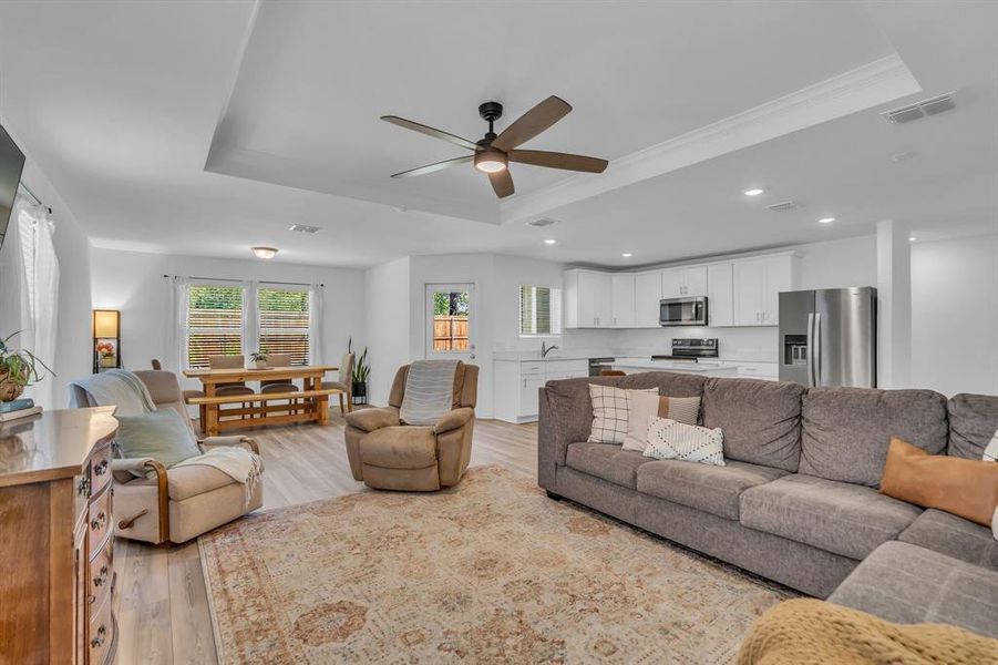 Living room with a raised ceiling, a ceiling fan, light wood finished floors, recessed lighting, and ornamental molding