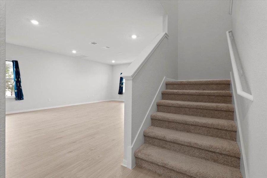 Carpeted staircase with white handrail, complemented by an expansive room featuring wood-finish flooring, recessed lighting, and a neutral wall color