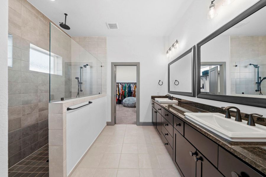 Spa-inspired primary bathroom featuring dual vanities with granite countertops, oversized mirrors, and modern lighting, complemented by a walk-in shower with sleek tile surround and direct access to a spacious walk-in closet.