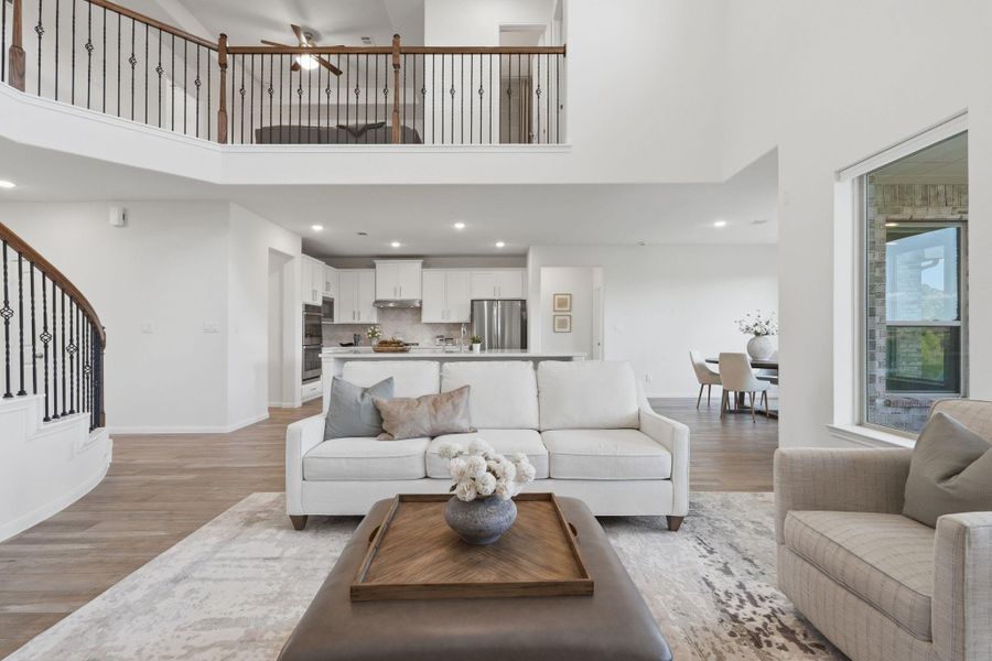 This photo showcases a spacious, open-concept living area with high ceilings and a modern design. The room features elegant wood tile flooring, a comfortable seating area, and a view into a sleek kitchen and dining space. A staircase with wrought iron railings leads to a second-floor balcony, adding to the airy, inviting atmosphere.