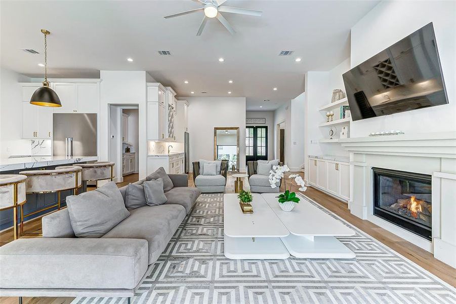 This spacious, modern living room features an open-concept design with a sleek kitchen, cozy fireplace, and large TV. Light tones and elegant furnishings create a warm, inviting atmosphere, perfect for entertaining or relaxing.