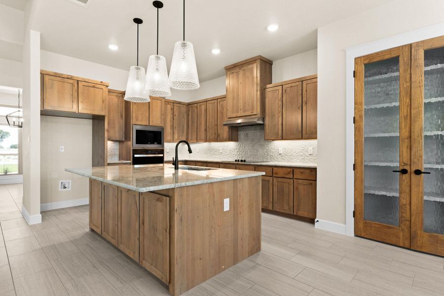 Kitchen with decorative light fixtures, brown cabinetry, light stone counters, backsplash, and wood finish floors Kitchen with decorative light fixtures, brown cabinetry, light stone counters, backsplash, and wood finish floors