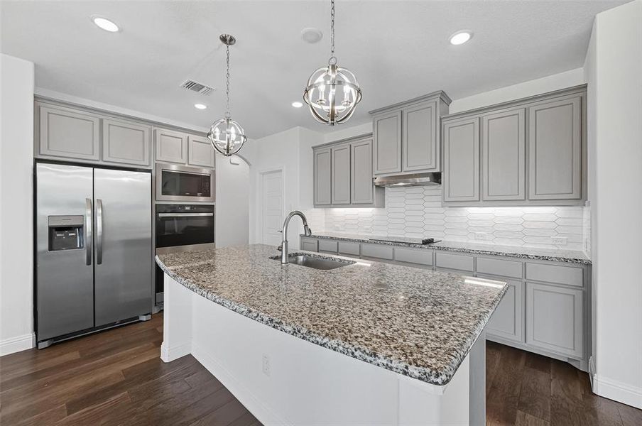 Kitchen with gray cabinetry, stainless steel appliances, light stone countertops, backsplash, and a center island with sink