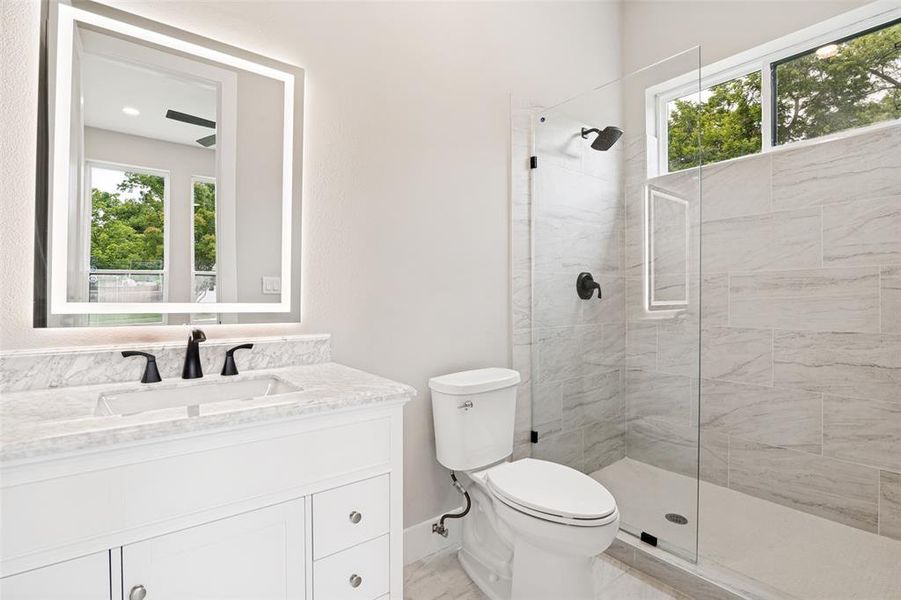 Bathroom featuring a white vanity with a stone countertop and integrated sink, an illuminated mirror, a glass-enclosed shower with matte black fixtures, and light-toned tile flooring and shower surround