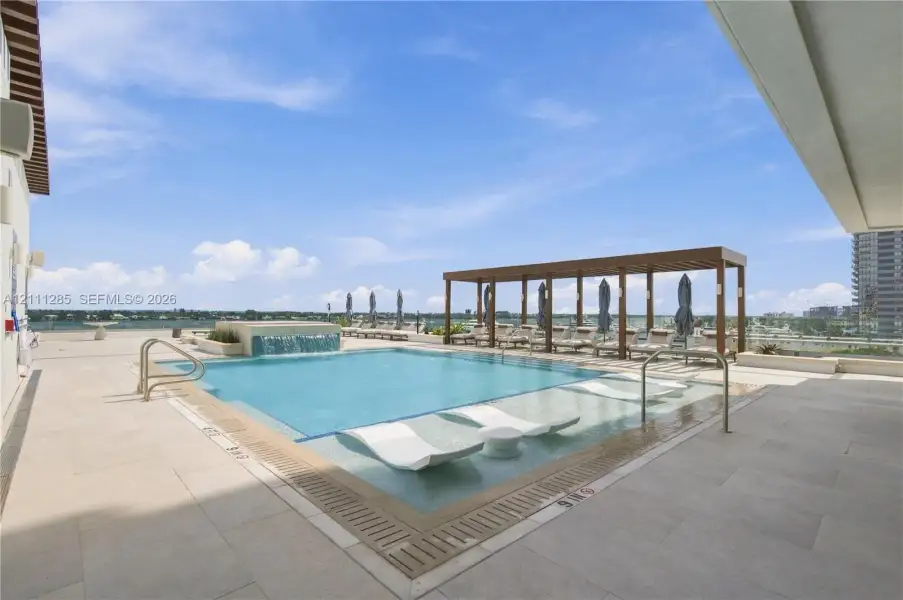 Rooftop pool featuring a water feature, baja shelf, large-format tile decking, and a wood-finish pergola Rooftop pool featuring a water feature, baja shelf, large-format tile decking, and a wood-finish pergola
