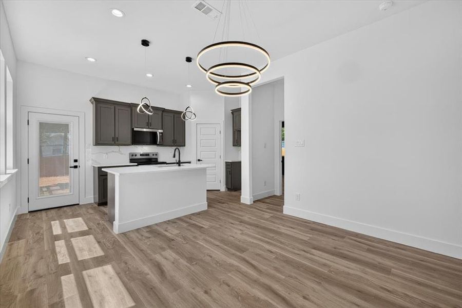 Open-concept area featuring wood-finish flooring, a kitchen island with a dark faucet, dark cabinetry, and a white tile backsplash Open-concept area featuring wood-finish flooring, a kitchen island with a dark faucet, dark cabinetry, and a white tile backsplash