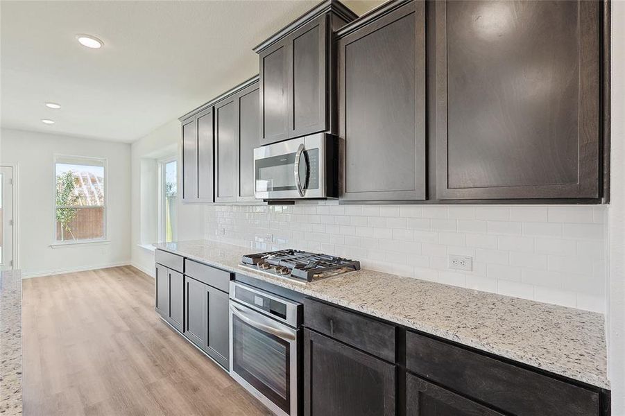 Kitchen featuring tasteful backsplash, light hardwood / wood-style flooring, light stone countertops, appliances with stainless steel finishes, and dark brown cabinetry