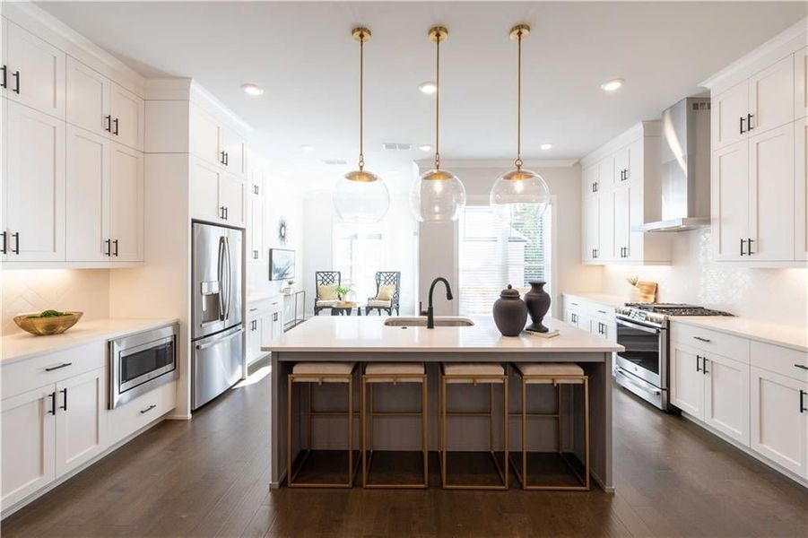 Kitchen with light countertops, hanging light fixtures, appliances with stainless steel finishes, wall chimney range hood, and an island with sink