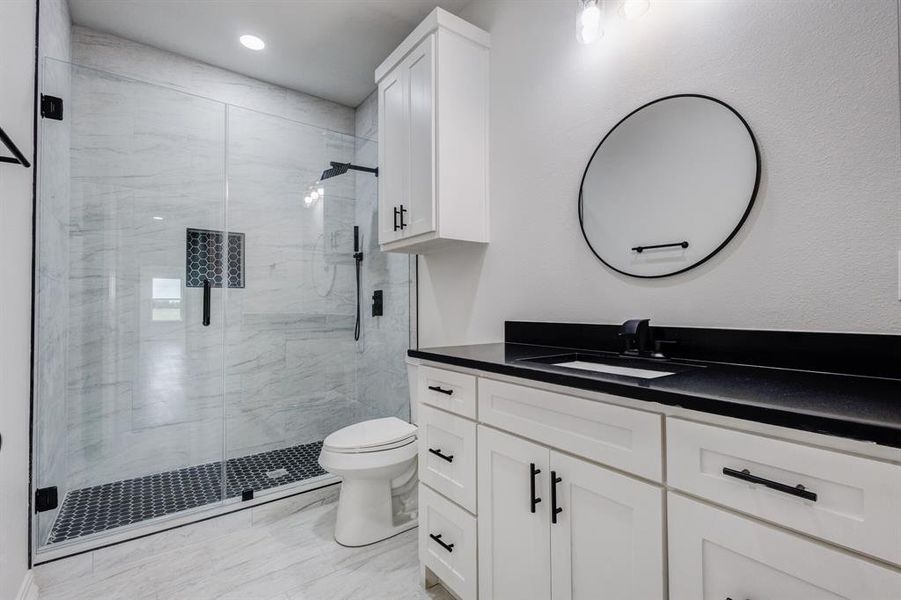Bathroom featuring a glass-enclosed shower with black hexagonal tile flooring, a white vanity with a dark countertop and undermount sink, and a circular mirror