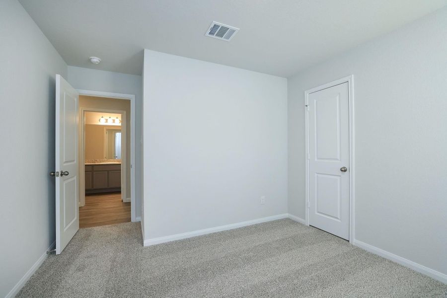 This is a bright, carpeted bedroom with neutral walls. It features a closet with a white door and an open doorway leading to a bathroom with vinyl plank flooring and modern lighting.