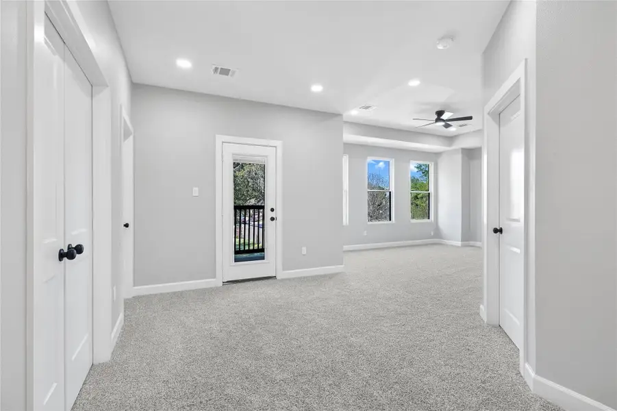 This spacious room features neutral gray walls and carpet, offering a bright and airy feel. It includes a ceiling fan, multiple windows for natural light, and a door leading to a small balcony, perfect for enjoying outdoor views.