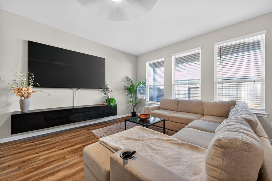 This inviting living room features a spacious beige sectional sofa, a large wall-mounted TV, and abundant natural light from three windows. The room is accented with plants and flowers, has warm wood flooring, and includes a sleek black entertainment console.