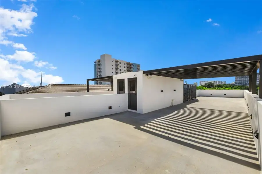 Rooftop terrace featuring concrete flooring, a partial louvered metal pergola, solid perimeter walls, and exterior wall-mounted lighting Rooftop terrace featuring concrete flooring, a partial louvered metal pergola, solid perimeter walls, and exterior wall-mounted lighting