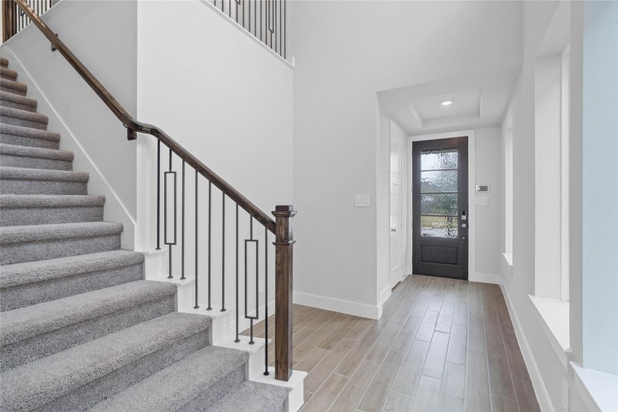 A bright and open interior entryway focuses on modern design and natural light. The staircase features iron rails and a rich, dark wood handrail, contrasting elegantly with the light carpeted steps. The space is amplified by high ceilings and generous natural light pouring in from large windows, creating an airy and welcoming first impression.