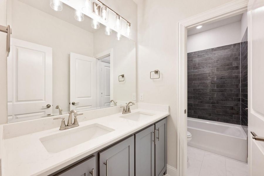 Bathroom with double sink vanity, gray cabinets, white countertop, and tub with dark tile surround