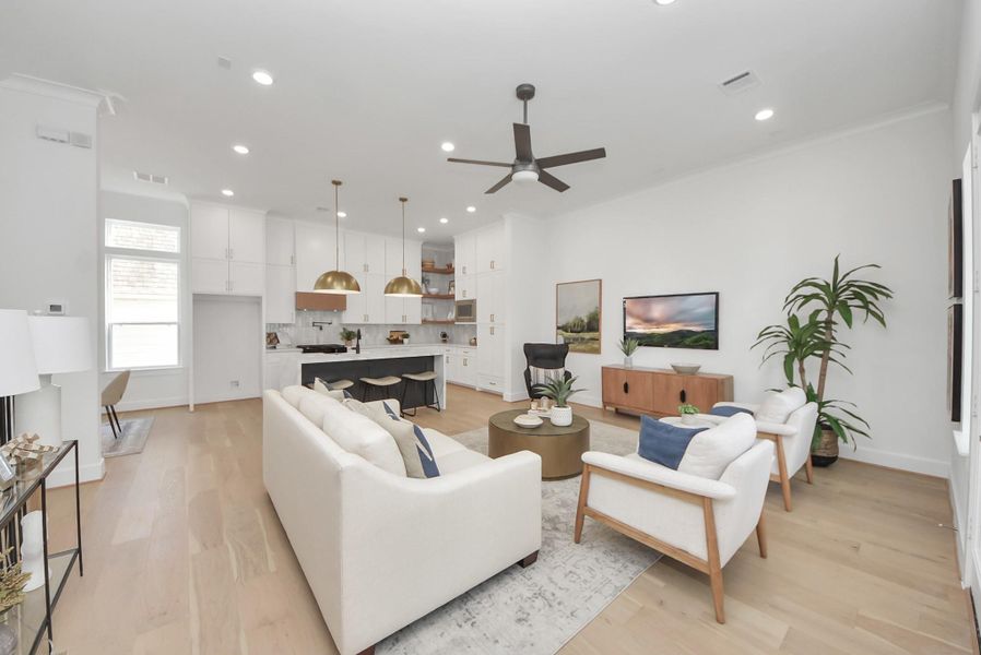 Welcome to this modern, open-concept living area, where light wood flooring and abundant natural light create a bright and inviting atmosphere. The sleek kitchen, with white cabinetry and gold pendant lights, flows seamlessly into a cozy living space featuring contemporary furniture and a ceiling fan—perfect for entertaining or relaxing with family and friends.