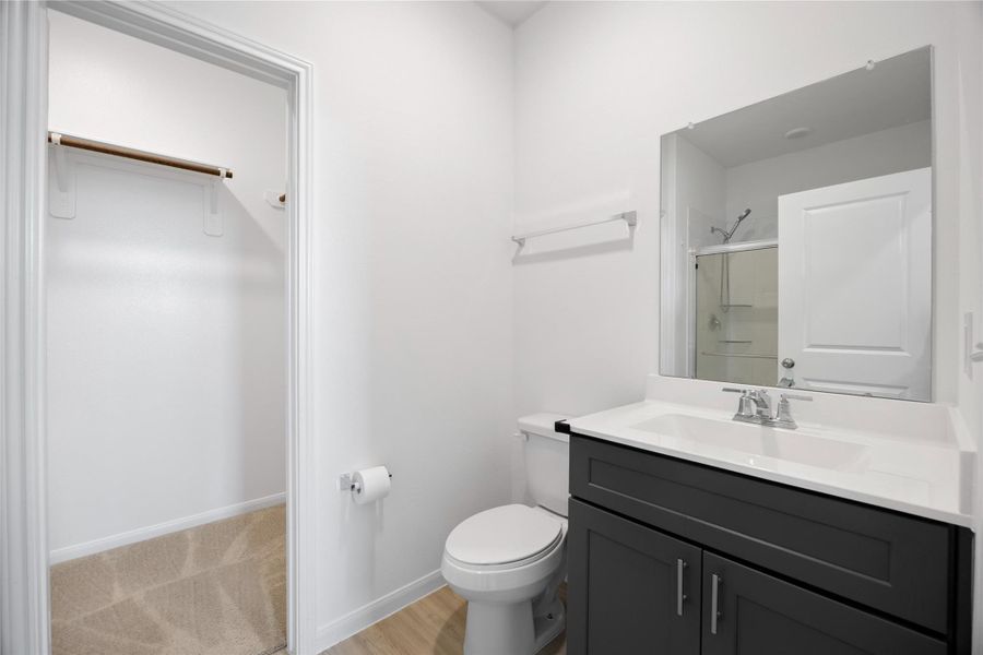 modern bathroom with a sleek vanity featuring a large mirror. It includes a toilet and a shower area with a glass door. Adjacent is a walk-in closet with ample storage space. The room is bright and clean with a neutral color palette.