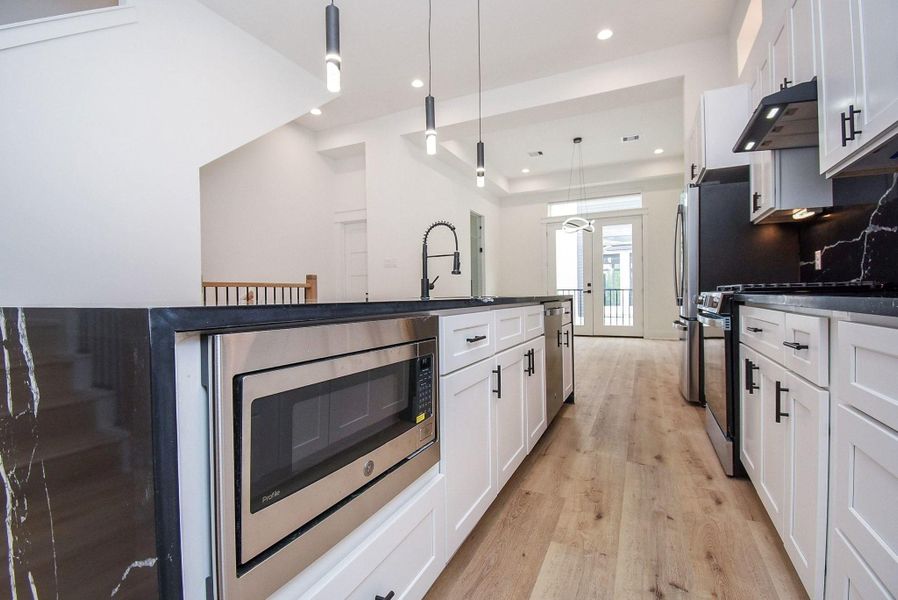 This modern kitchen features sleek white cabinetry, black countertops, and stainless steel appliances, including a built-in microwave. The space is brightened by ample natural light and contemporary pendant lighting, with wood flooring adding warmth.