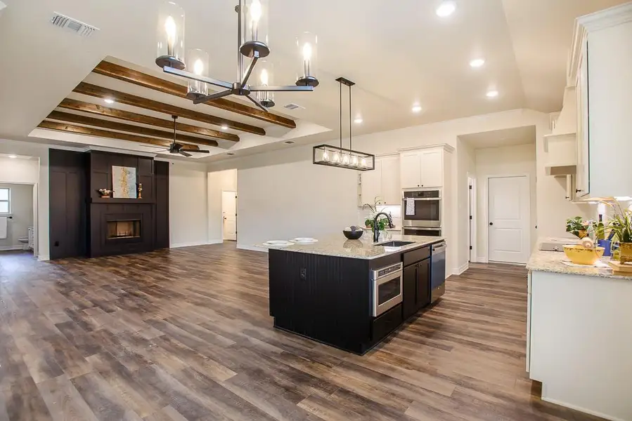 Kitchen with ceiling fan, a large fireplace, dark wood-style floors, white cabinetry, and a kitchen island with sink Kitchen with ceiling fan, a large fireplace, dark wood-style floors, white cabinetry, and a kitchen island with sink