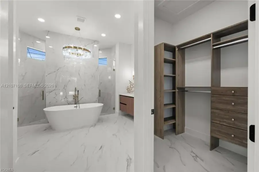 Freestanding soaking tub with floor-mounted faucet, floor-to-ceiling stone-style wall cladding, crystal chandelier, and walk-in closet featuring built-in wood-finish shelving and cabinetry