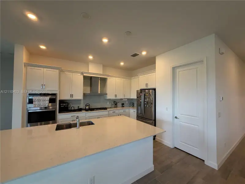 Kitchen featuring a large island with quartz countertops, custom white cabinetry, stainless steel double wall ovens, built-in gas cooktop with chimney-style range hood, and wood-finish flooring