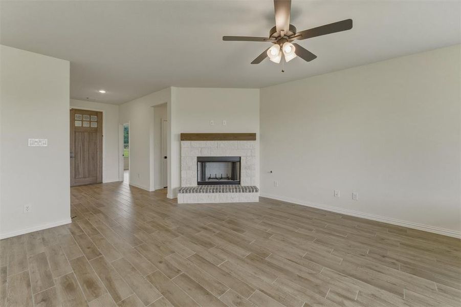 Spacious living area featuring light wood-finish flooring, a contemporary fireplace with a stone surround and wood mantel, a ceiling fan with integrated lighting, and a wood-paneled front door with clear glass inserts Spacious living area featuring light wood-finish flooring, a contemporary fireplace with a stone surround and wood mantel, a ceiling fan with integrated lighting, and a wood-paneled front door with clear glass inserts