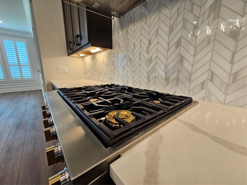 This kitchen features a modern gas stove with a sleek, stainless steel finish and gold accents. It has a stylish herringbone tile backsplash and elegant quartz countertops. The space receives natural light from nearby windows with plantation shutters, highlighting the rich wood flooring. This kitchen features a modern gas stove with a sleek, stainless steel finish and gold accents. It has a stylish herringbone tile backsplash and elegant quartz countertops. The space receives natural light from nearby windows with plantation shutters, highlighting the rich wood flooring.