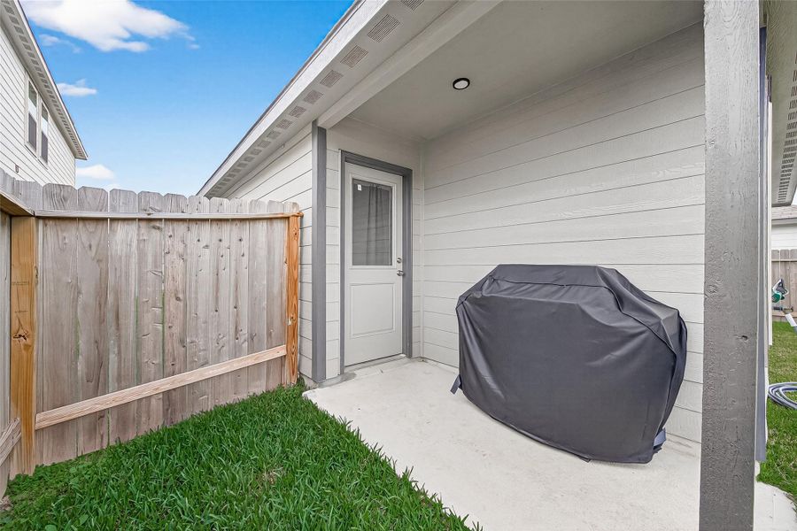 This photo shows a small, covered patio area with a barbecue grill and green lawn. The space is partially enclosed by a wooden fence, offering privacy and a cozy outdoor area.