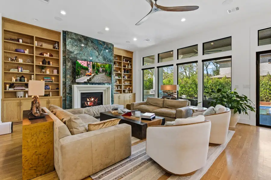 Inviting living room centered around a statement fireplace with designer surround and custom built-in shelving. Wide-plank hardwood flooring, recessed lighting, and expansive windows fill the space with natural light, while the open layout creates a seamless connection to the kitchen and outdoor living areas.