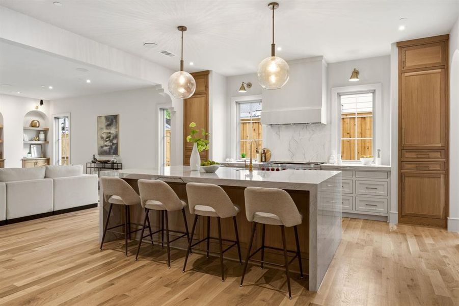 Spacious kitchen featuring a large island with seating, pendant lighting, built-in cabinetry, wood-finish flooring, and a stone backsplash Spacious kitchen featuring a large island with seating, pendant lighting, built-in cabinetry, wood-finish flooring, and a stone backsplash