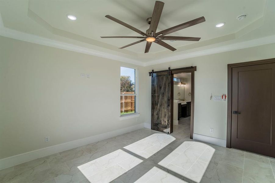 Bedroom with a barn door, a raised ceiling, ceiling fan, and connected bathroom Bedroom with a barn door, a raised ceiling, ceiling fan, and connected bathroom