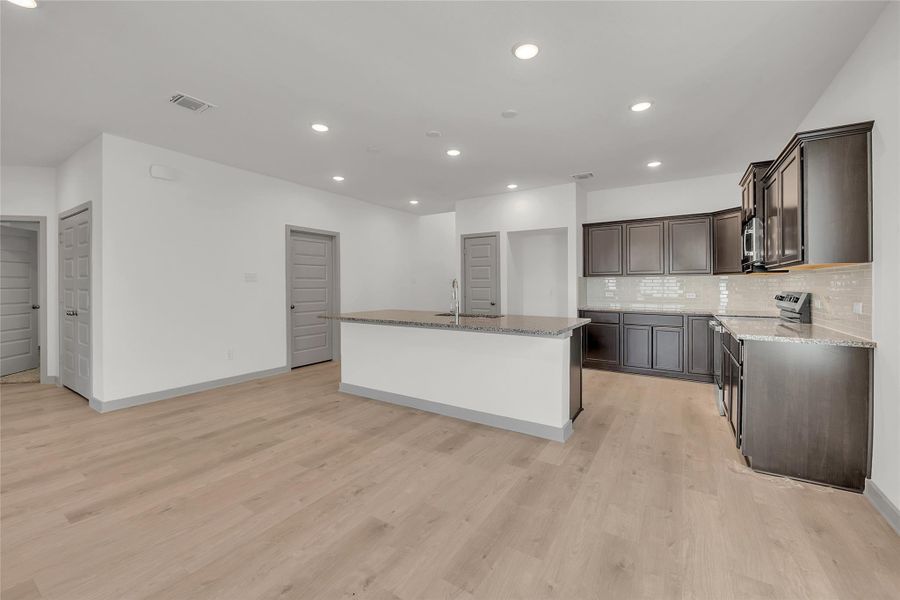 Kitchen featuring decorative backsplash, recessed lighting, light stone counters, a kitchen island with sink, and dark brown cabinetry