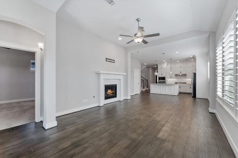 This is a spacious, well-lit living room featuring laminate wood floors, a white fireplace, large windows with plantation shutters, and a ceiling fan. The room has a clean, modern feel with neutral colors.