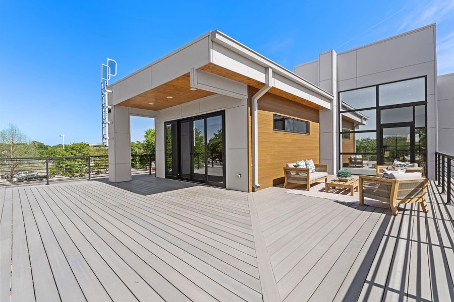 The rooftop deck is one of this home's most unexpected and compelling features — a sprawling decked outdoor living platform with a dedicated covered section finished in a warm wood soffit ceiling and recessed lighting, connected to the interior through large sliding glass panels. Black horizontal cable railings frame the full perimeter, and the open expanse beyond the covered portion gives the space room to breathe under open sky.