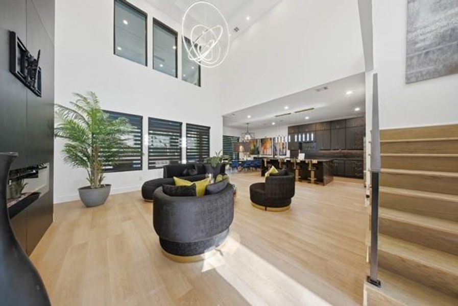 Open-concept main living area with high ceilings, blonde wood-finish flooring, a modern horizontal bar staircase, built-in bar with pendant lighting, and large windows with horizontal blinds