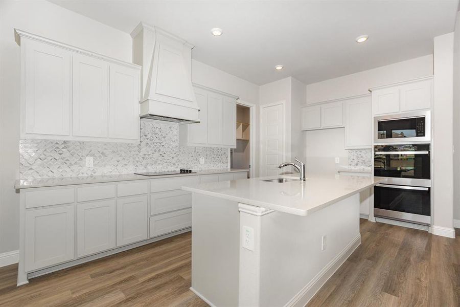 Kitchen featuring backsplash, double oven, light wood-style floors, a center island with sink, and light stone counters