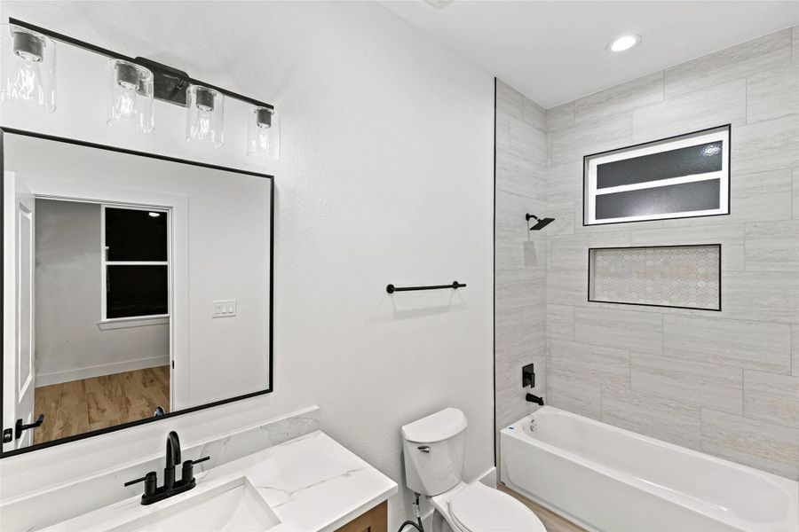 Modern bathroom featuring a sleek vanity with a black faucet, a large framed mirror, and a stylish light fixture. The bathtub area is accented with neutral tiles and a built-in niche, complemented by a window for natural light. Modern bathroom featuring a sleek vanity with a black faucet, a large framed mirror, and a stylish light fixture. The bathtub area is accented with neutral tiles and a built-in niche, complemented by a window for natural light.