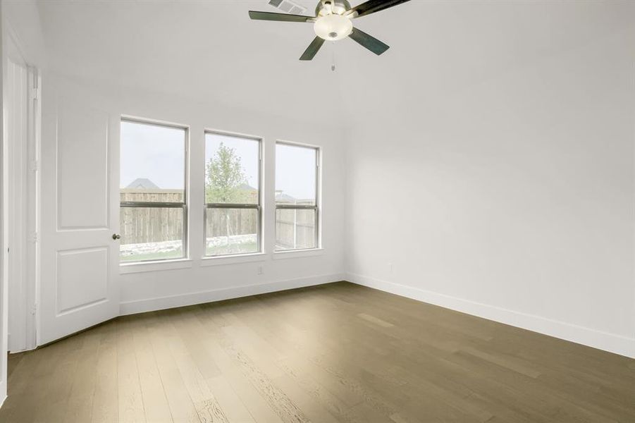 Spacious room featuring light wood-finish flooring, three large windows, a white paneled door, and a ceiling fan with integrated lighting