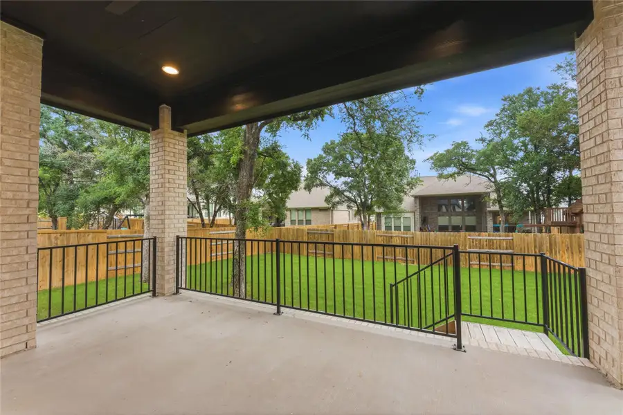 Covered patio with recessed lighting, brick columns, concrete flooring, and black metal railings, overlooking a fenced backyard with a manicured lawn