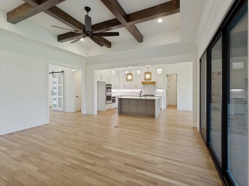 Open-concept living area featuring light wood-finish flooring, a coffered ceiling with dark wood beams, and a black ceiling fan Open-concept living area featuring light wood-finish flooring, a coffered ceiling with dark wood beams, and a black ceiling fan
