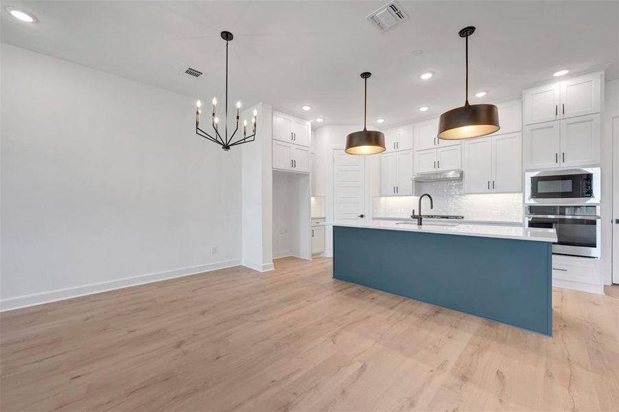 Open-concept kitchen and dining area featuring light wood-finish flooring, a contemporary black chandelier, a central island with a blue base and white countertop, white shaker cabinetry, and stainless steel appliances Open-concept kitchen and dining area featuring light wood-finish flooring, a contemporary black chandelier, a central island with a blue base and white countertop, white shaker cabinetry, and stainless steel appliances