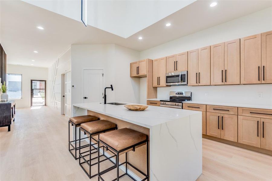 Kitchen featuring light brown cabinets, light hardwood / wood-style flooring, light stone countertops, an island with sink, and stainless steel appliances