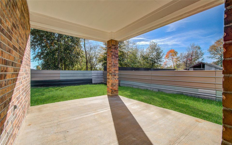 This photo shows a covered patio with a concrete floor and brick columns, leading to a grassy backyard. A metal fence encloses the area, with trees visible beyond the fence, providing a sense of privacy and outdoor space.
