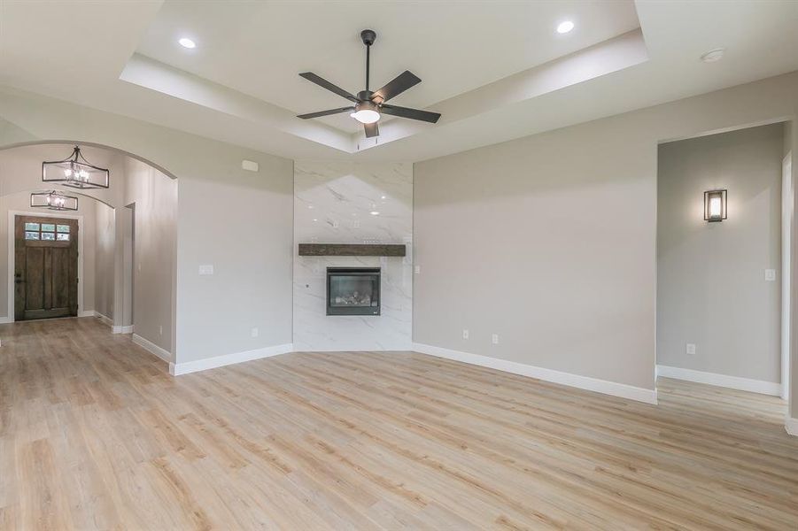 Unfurnished living room with a tray ceiling, arched walkways, ceiling fan, light wood-style floors, and a premium fireplace Unfurnished living room with a tray ceiling, arched walkways, ceiling fan, light wood-style floors, and a premium fireplace