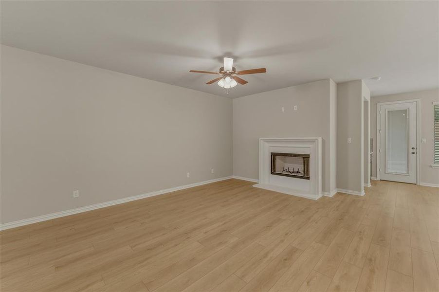 Spacious room featuring light wood-finish flooring, a white mantel fireplace with a dark insert, a ceiling fan with light fixture, and neutral wall tones