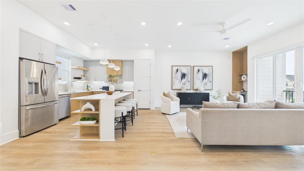 This photo showcases a bright, open-concept living area featuring a modern kitchen with stainless steel appliances and an island with seating. The living space has large windows, sleek furnishings, and light wood flooring, providing a contemporary and welcoming atmosphere.