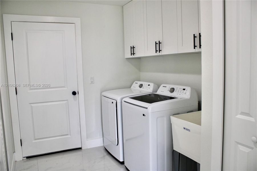 Laundry room featuring white shaker-style wall cabinets with matte black hardware, a utility sink, and large-format tile flooring