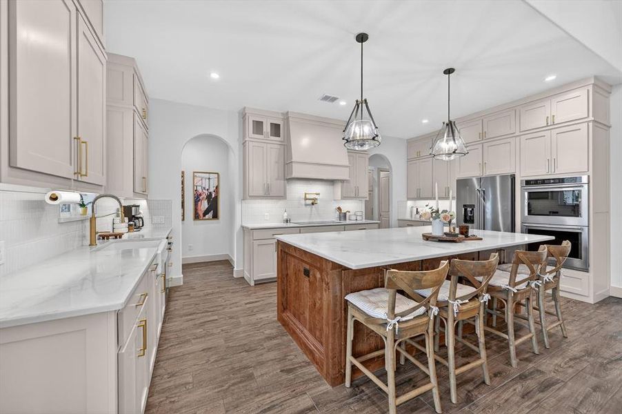 Expansive kitchen featuring a large central island with a white countertop, extensive light-toned cabinetry, stainless steel appliances, wood-finish flooring, and pendant lighting