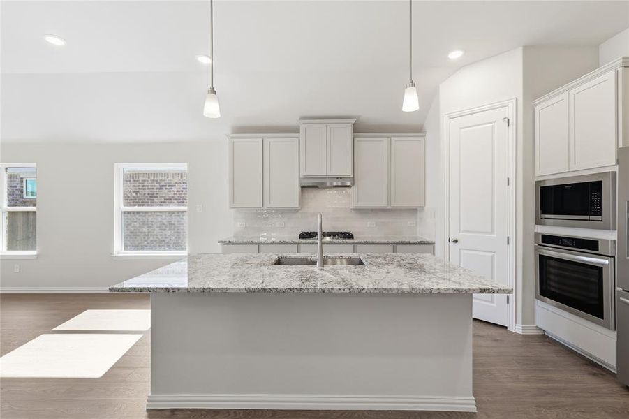 Kitchen featuring dark wood-style flooring, light stone countertops, stainless steel appliances, a kitchen island with sink, and pendant lighting Kitchen featuring dark wood-style flooring, light stone countertops, stainless steel appliances, a kitchen island with sink, and pendant lighting