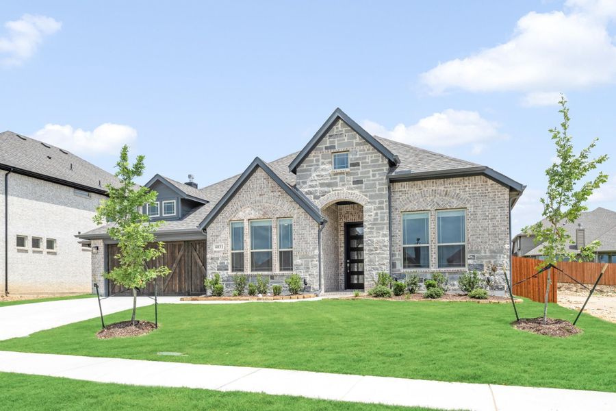 Single-story stone and brick home with arched entry, wood garage door, and manicured front lawn with young trees. Single-story stone and brick home with arched entry, wood garage door, and manicured front lawn with young trees.
